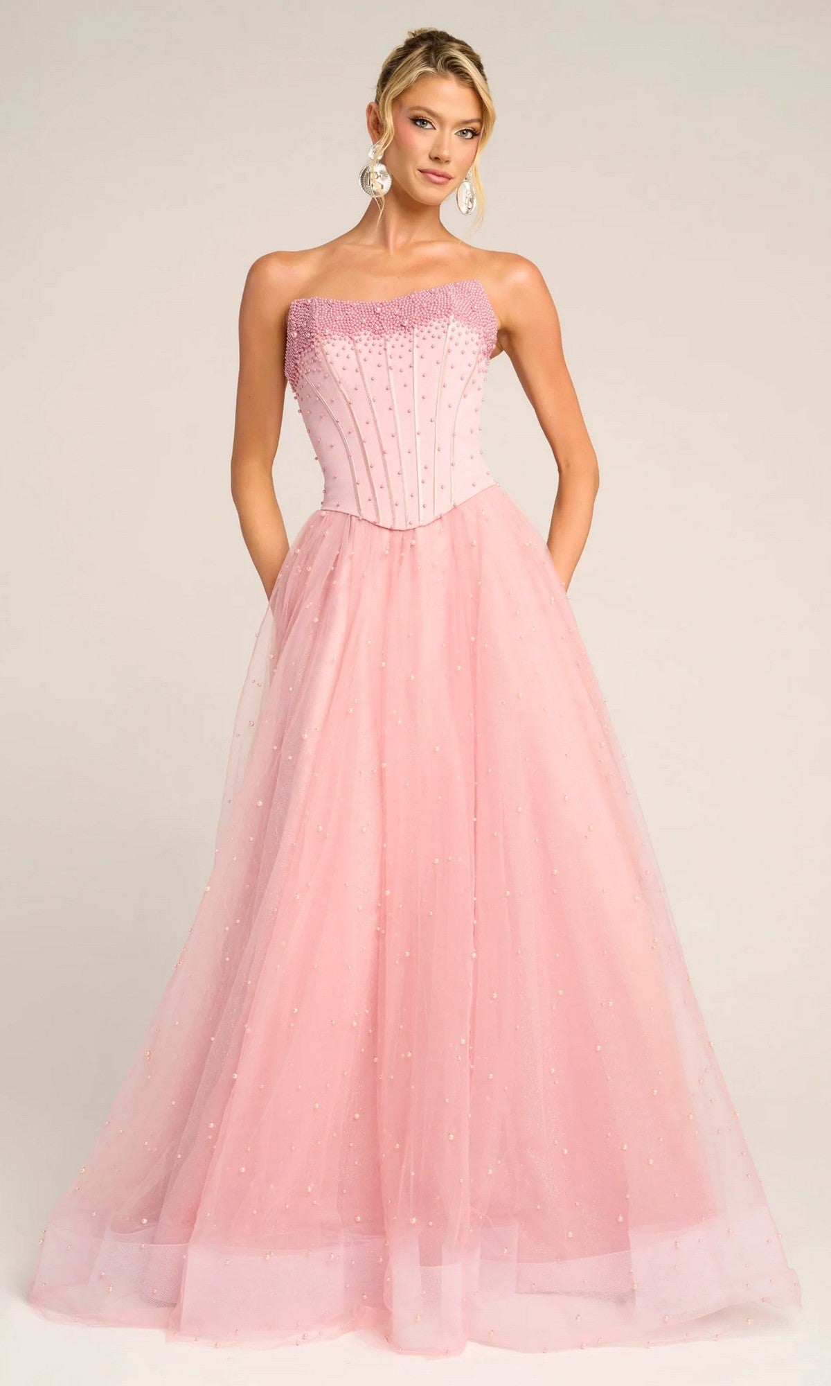 A woman poses in the Portia and Scarlett PS26034 long formal dress in pink, featuring a strapless beaded corset bodice and a flowing glitter tulle skirt.